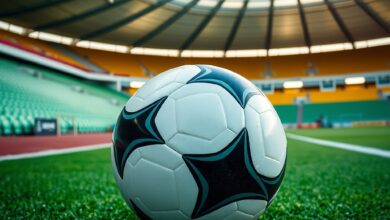Bola de futebol sobre relvado verde num estádio moderno sob holofotes desportivos em dia de jogo.